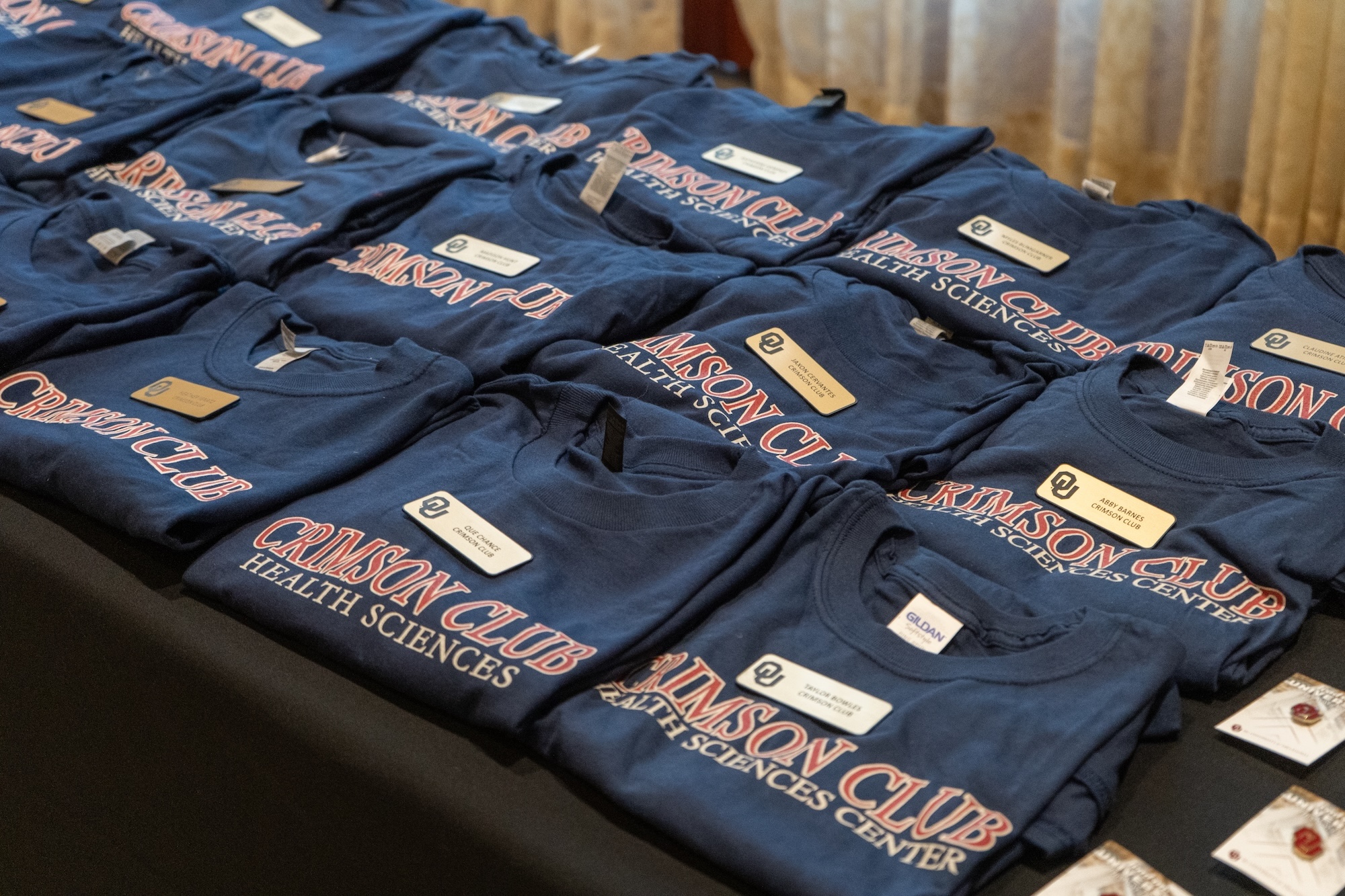 A picture of a table of Crimson Clubs t-shirts with a golden name tag on each shirt.