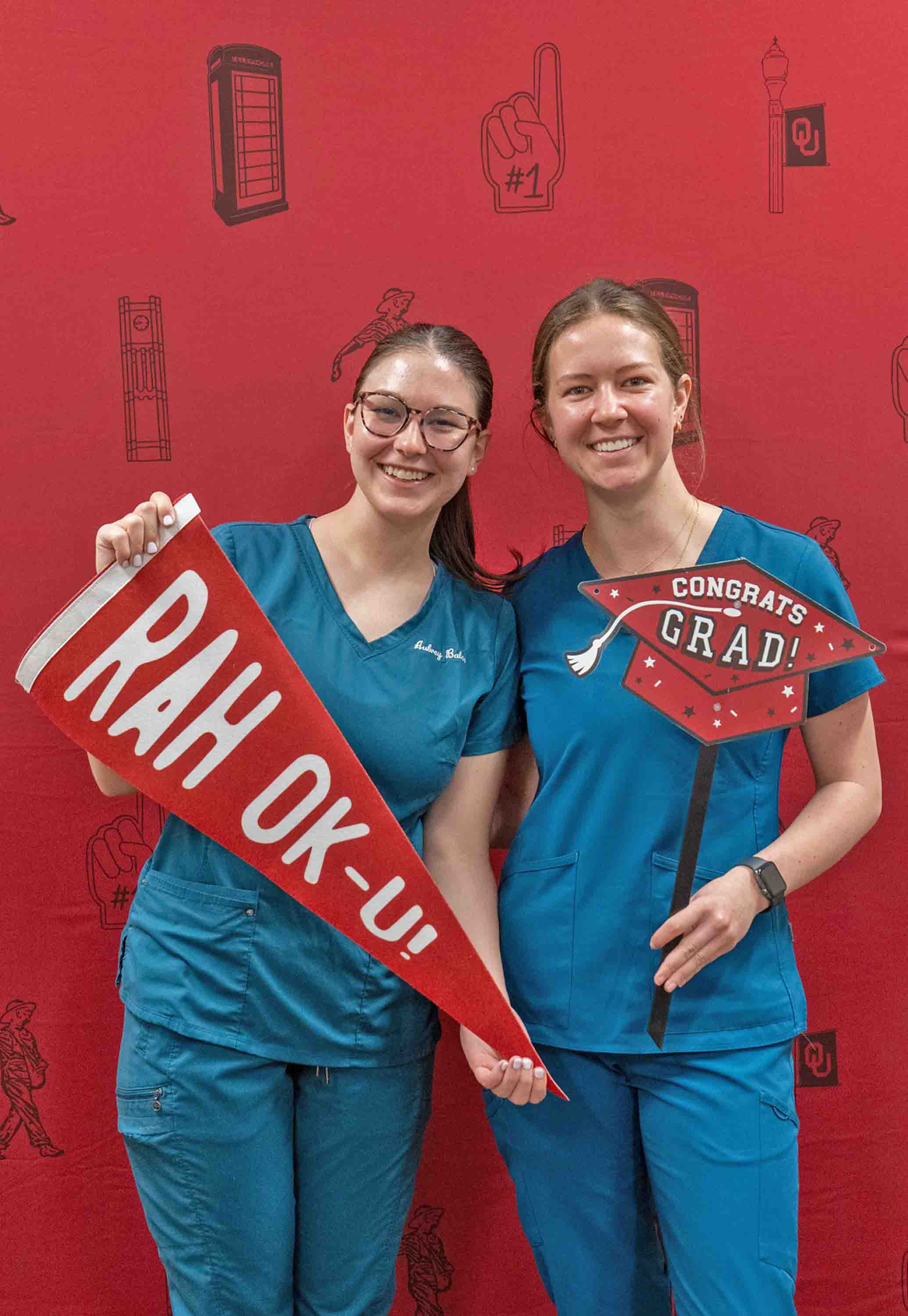 Students posing for a photo in front of a crimson background during the Graduation Gear-Up event. One student is holding a 'Rah OK-U pennant and the other student is holding a 'Congrats Grad' sign shaped like a graduation cap.