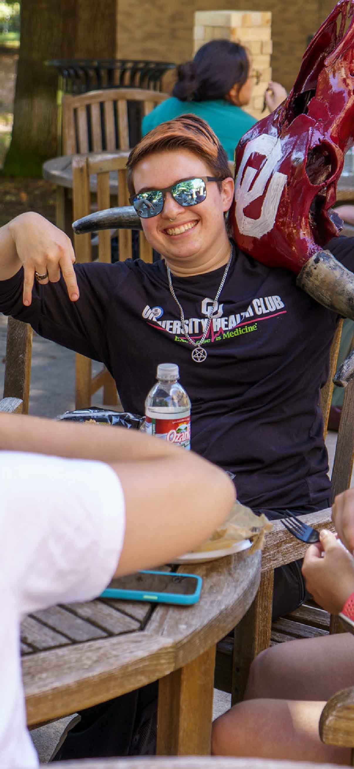 A student sits with sunglasses on while holding an upside-down longhorn skull on her left shoulder during Bevo BBQ. The student also signs 'horns down' with her right hand.