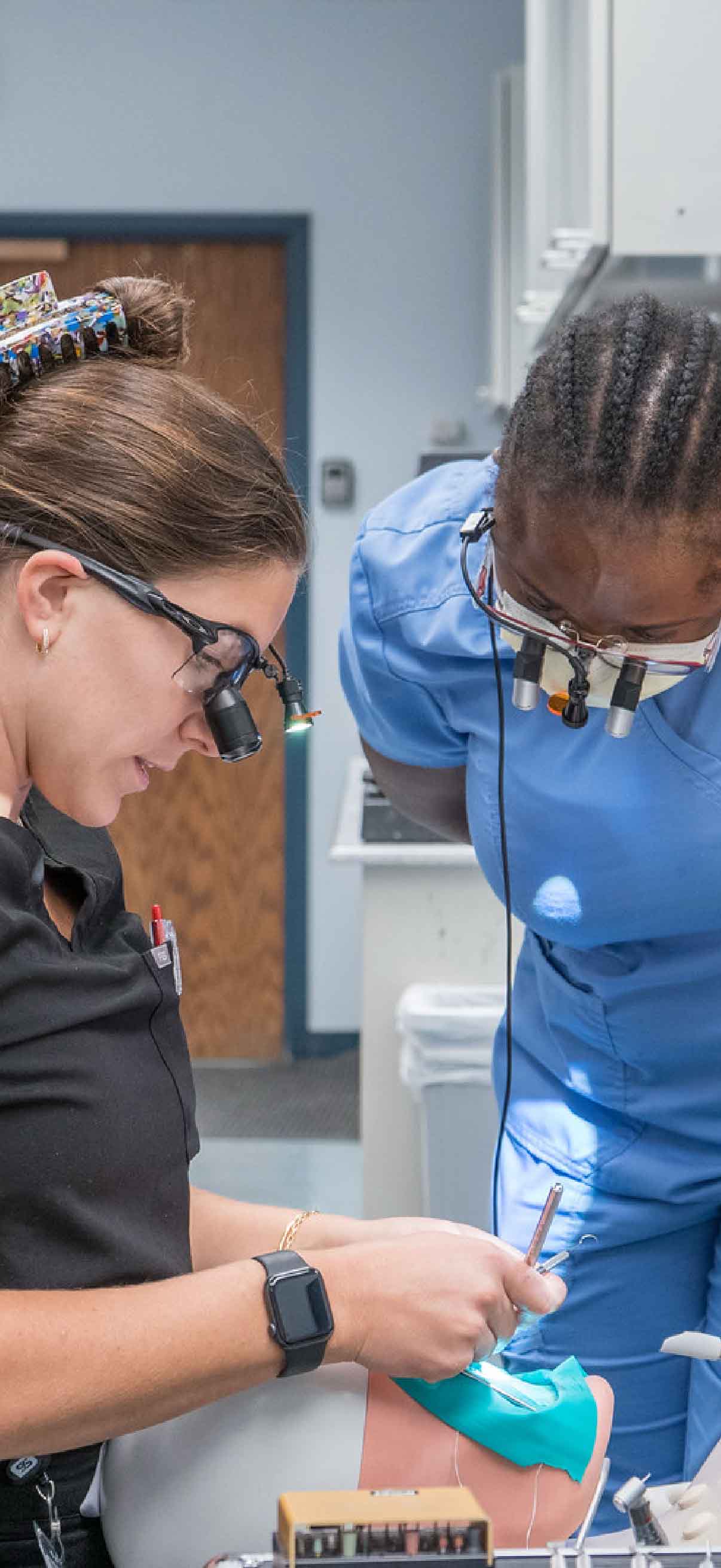 Two dental students work on model teeth in a lab. One student is sitting and flossing the teeth while the other student stands and observes.