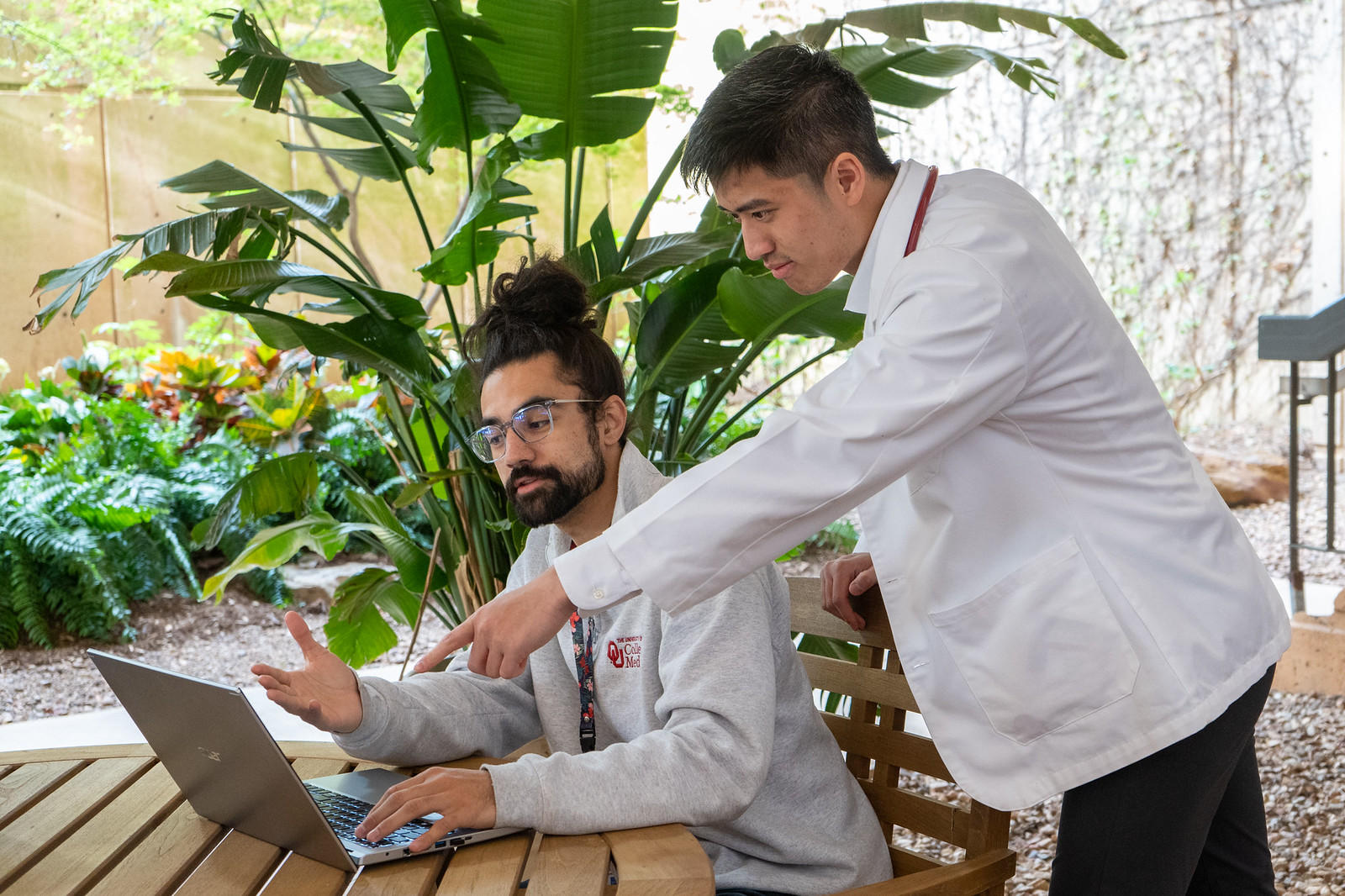 two students sitting on wooden chairs outside the College of Allied Health looking at a computer screen.